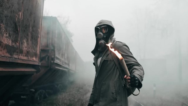 Hooded man in protective gas mask standing by abandoned carriages at destroyed train station holding burning fire in hand. Apocalypse, survivor after nuclear or chemical disaster. Extreme slow motion