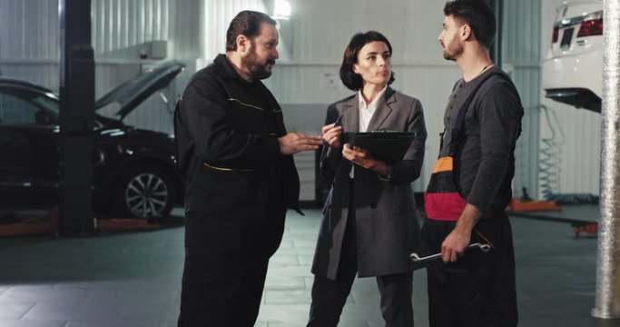 In a modern auto service center office worker lady in a suit have a conversation with the main mechanic and other young mechanic they analyzing the plan of work