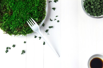 A plastic plate with green moss stands on a white wooden table and next to it are containers with dried parsley and soy sauce. A disposable fork lies on the moss.