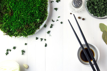 A plastic plate with green moss stands on a white wooden table and next to it are containers with dried parsley soy sauce 