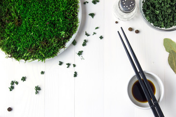 A plastic plate with green moss stands on a white wooden table and next to it are containers with dried parsley soy sauce and a salt shaker. And on the side are black chopsticks.