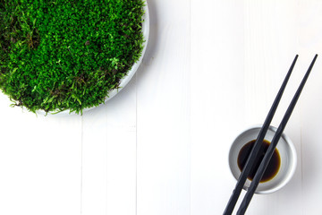 A plastic plate with green moss stands on a white wooden table and next to it is a bowl of soy sauce on which are chopsticks.