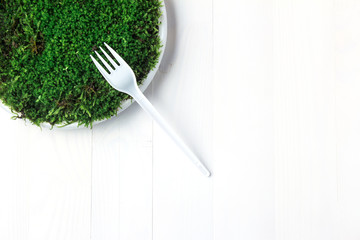 A plastic plate with green moss stands on a white wooden table and on it lies a disposable fork.
