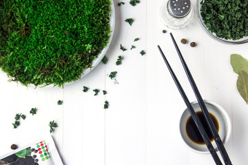 A round plastic plate with green moss stands on a white wooden table and next to it is scattered dried parsley and black chopsticks in a bowl with soy sauce.