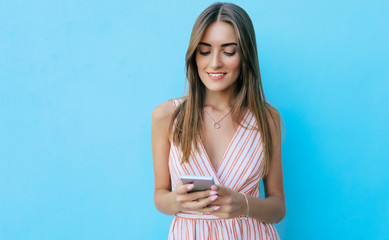 Facebook fan. Mesmerizing woman in a striped summer dress is posing with a smartphone in her hands, smiling while reading something on her phone.