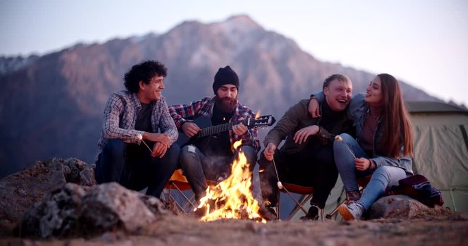 Group of four hipster frends relaxing on top of a mountain, sitting near bonfire, roasting marshmallows and enjoying their time - friendship, youth, love concept 4k footage