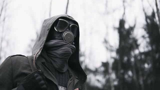 Wandering human in military clothes and protective gas mask walking in dark forest. Stalker concept, portrait of survivor after nuclear or chemical war. Post apocalyptic world. Stedicam following shot