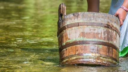 A young girl is washing a white cloth in a wooden bucket. She is standing in the water of the river.