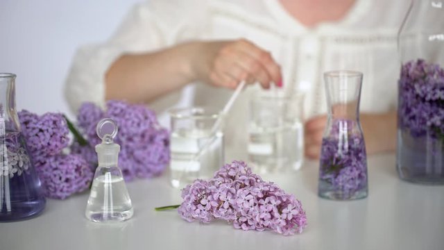 Making body care production beauty aroma perfume lilac flowers background on table, Hand of female perfumer lab technician chemist cosmetician pouring cosmetic aroma glass test tube oil in liquid, 4 K