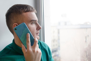 Serious pensive guy is talking on his cell mobile phone, young man having conversation on smartphone and looking at window at home. Male thinking dreaming and looking out the window.