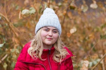 Medium horizontal photo of pretty smiling young woman with long blond hair wearing red puffy warm coat and knitted hat staring with dreamy expression in front of late fall landscape