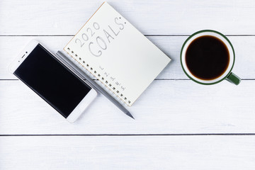 2020 new year goals, plan, action text on notepad on white wooden boards. 2020 goals on blank note paper with copy space for text and smartphone. Cup of coffee over wooden boards