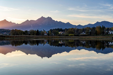 Peaceful scene of beautiful autumn mountain landscape with small settlement, calm lake, colorful trees and high peaks in High Tatras, Slovakia.