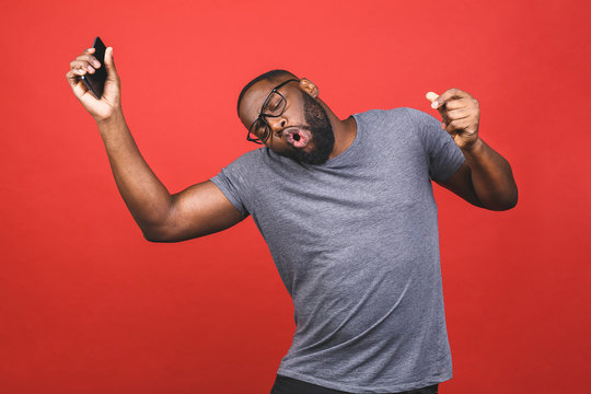 Indoor Portrait Of Young Handsome African Man Isolated On Red Background During Dance To Playlist He Is Listening From Cellphone, Being Excited.