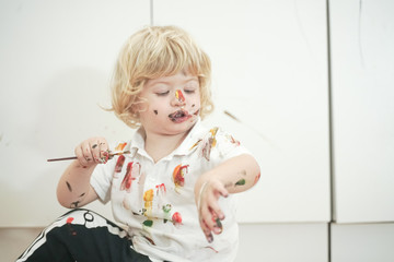 two year old boy with hands and face painted in colorful paints ready for more fun. dirty and happy kid sitting on the floor in white room background.