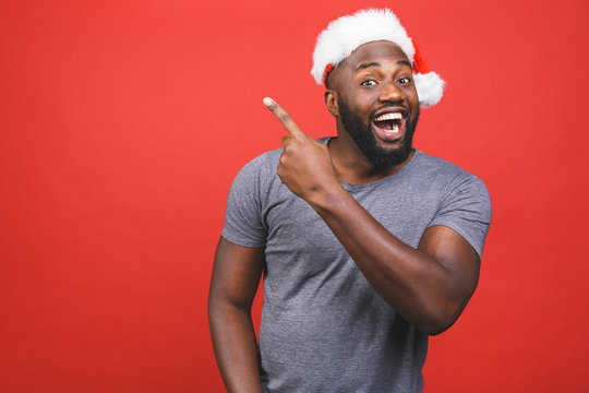 Best price for you, look here concept. Handsome african man in red christmas cap pointing fingers and looking at camera isolated over red background. Christmas sale and black friday.