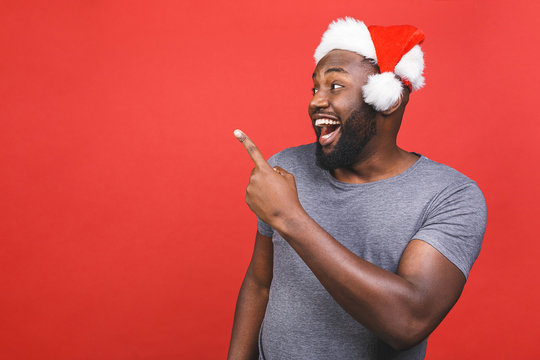 Best price for you, look here concept. Handsome african man in red christmas cap pointing fingers and looking aside isolated over red background. Christmas sale and black friday.