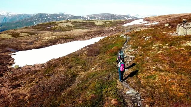 A drone flight following a couple carrying big backpack hikes through dry trail, with snow patches in few paces. High mountains in the back covered with snow. Beautiful day for a trek.