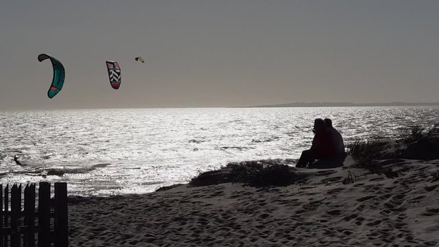 Kitesurfer jump, catch airtime in Cape Town beach. Romantic silhouettes sitting on sand dune at silver beach sunset. Sunlight reflect on choppy ocean waves. Kitesurfing on glistening sea waves.