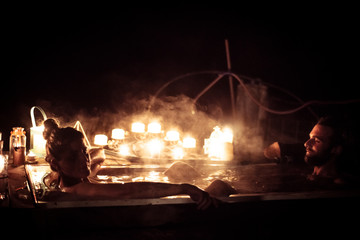 young couple sit in hot tub outdoors at night with romantic candles