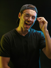 Emotional studio portrait of a young attractive man in a cap and glasses on a dark background