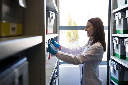 Side view of woman in white robe putting box with samples on shelf in lab storage