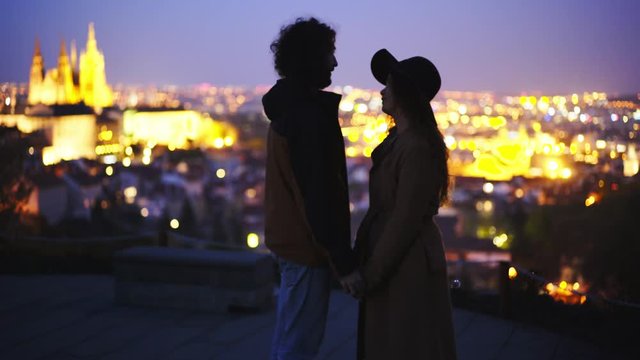 Silhouette couple enjoying togetherness against cityscape. Medium shot young girl and man on hill top above Prague. Side view lovers holding hands and kissing at background illuminated city 