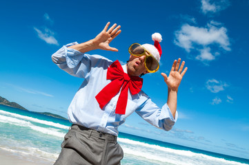 Cheerful businessman in Santa hat, sunglasses and big red Christmas bow dancing on a tropical beach