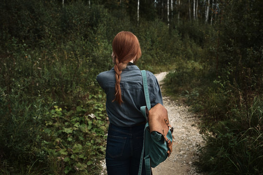 Close Frame. A Girl With A Red Braid In A Denim Shirt With A Backpack Walks Along A Path In The Middle Of The Forest, A Frame From The Back.