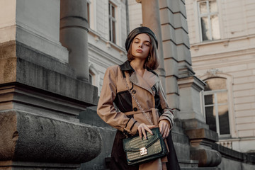 Outdoor autumn fashion portrait of young elegant woman wearing beige black trench coat, beret, with green faux leather textured crocodile shoulder bag, posing in street. Copy, empty space for text - obrazy, fototapety, plakaty