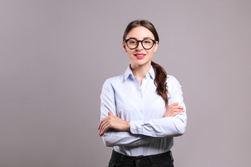 Young beautiful confident businesswoman wearing blue blouse & black frame glasses, standing w/ folded hands & smiling. Attractive brunette female with crossed arms. Background, close up, copy space.