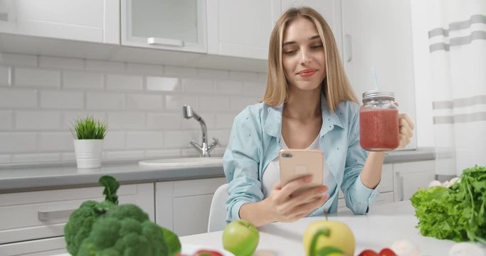 Pretty young blonde lady drinking fresh homemade smothie and scrolling smartphone sitting at table in kitchen, lifestyle