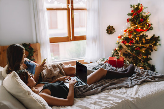 Two women friends in winter holidays at home looking something on laptop near christmas tree in cozy interior. Interior with christmas decorations. - Powered by Adobe