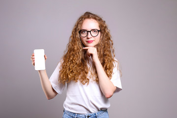 Young beautiful woman with long curly hair posing over isolated grey background & holding smartphone. Portrait of teenage female model wearing white t-shirt, showing emotions. Close up, copy space.