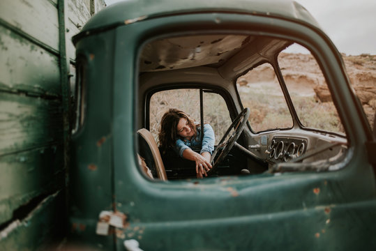 Thoughtful young woman in shorts and denim shirt standing leaning in a windows of a green truck with wooden body with closed eyes on nature background