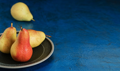 Three ripe pears in droplets of water in a ceramic plate on a blue background. Organic fruits close up. Space for text. Fresh seasonal produce. Agriculture and gardening, fresh harvest, concept.