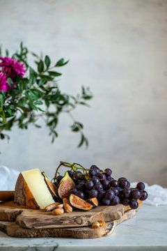 Sliced ripe figs and dark blue grapes next to piece of cheese on wooden cutting boards near bouquet of pink flowers among green leaves against blurred gray wall