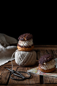 Homemade choux pastries with cream and chocolate scissors and ball of yarn arranged on texture wooden surface against black background