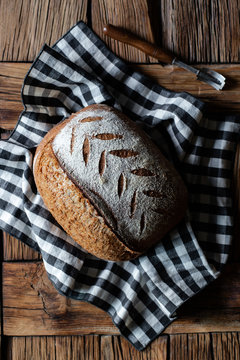 Top view of loaf of fresh spelt bread placed on checkered napkin near tool on lumber table