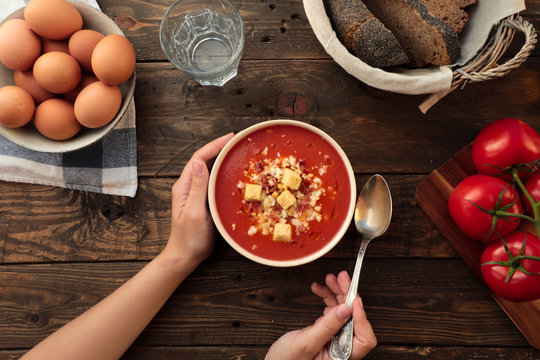 From above shot of unrecognizable person holding a bowl of red gazpacho in a rustic table with egg, bread, and fresh tomatoes arrangements