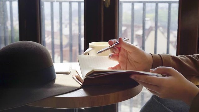  Woman drinking coffee near the window.View cropped hands holding pen and writing ,tourist planning our trip or student prepare for the exam, making to do list in notepad while sitting in cafe 