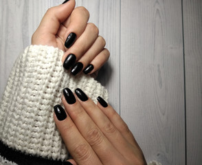 Coal-black nail coating with lamp highlights on delicate female hands in a white sweater. Black gel...