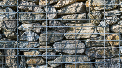 Gabion retaining wall texture. Closeup of stacked stones in wire mesh cage. Background of protective fence detail made from sunlit gray pieces of rocks in a metal grid with shadows on boulder masonry.