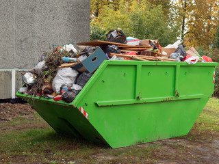 Big green color metal skip container full of tras by a house building.