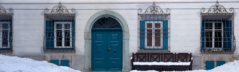 Fototapeta na wymiar ancient house facade with blue door, with wooden bench and snow drifts in front
