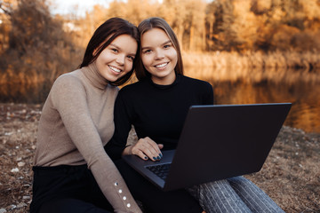 Cheerful two young twin sisters in casual outfit with bright smile use notebook or laptop looking in camera at the park.