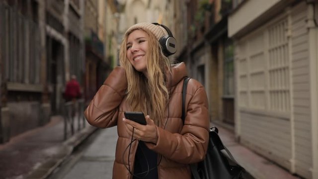 Smiling and laughing handsome young woman girl listening music from his smartphone in headphones, dancing on street of old town center in europe day. Slow motion