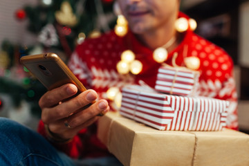 Christmas shopping online. Man buying New year presents using smartphone. Guy holding gift boxes and phone by tree