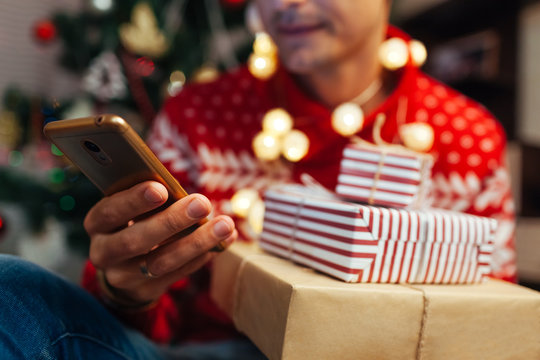 Christmas shopping online. Man buying New year presents using smartphone. Guy holding gift boxes and phone by tree