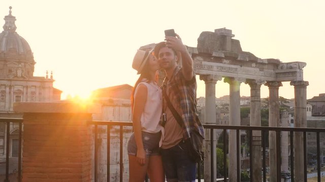 Young couple tourist looking at Roman Forum at sunrise and kissing taking selfie. Historical imperial Foro Romano in Rome, Italy from panoramic point of view.